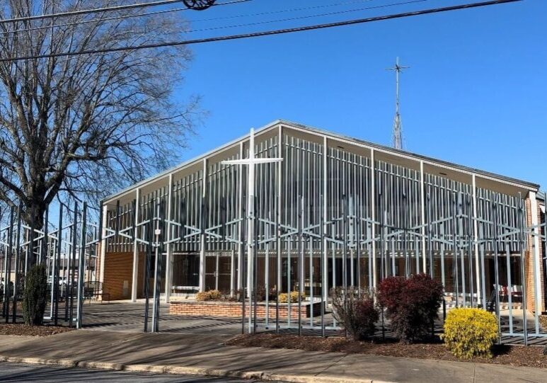 A modern church building with large glass windows, metal fence, and a cross on top, surrounded by landscaped shrubs and trees, under a clear blue sky.