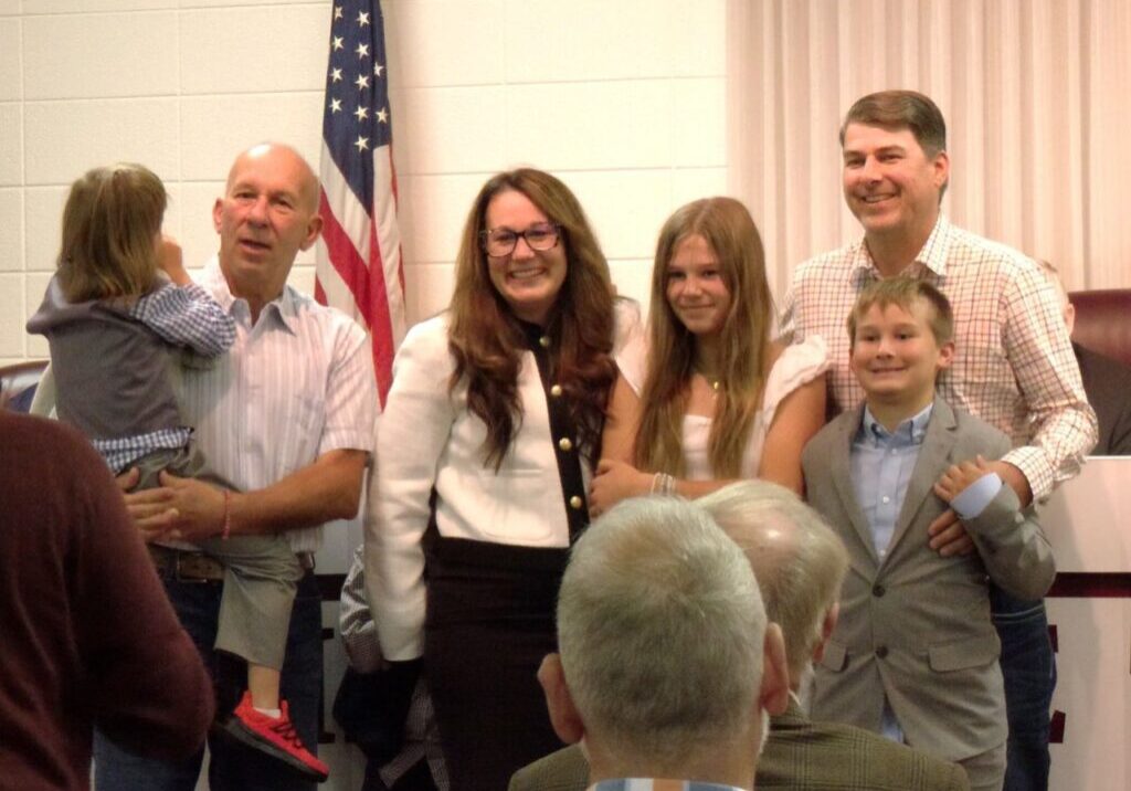 A group of five people, including three adults and two children, poses together in a room with an American flag in the background.