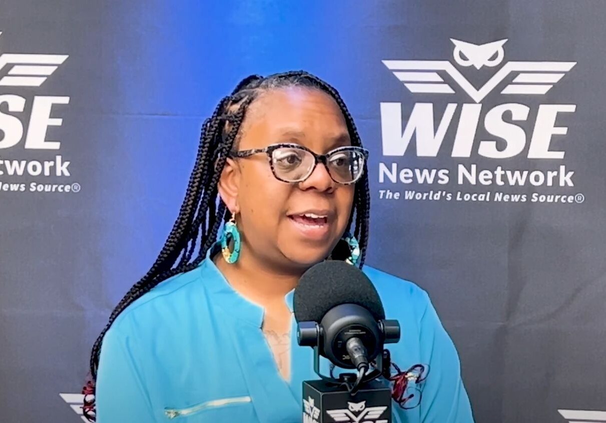 A woman with long hair and glasses is talking into a microphone.