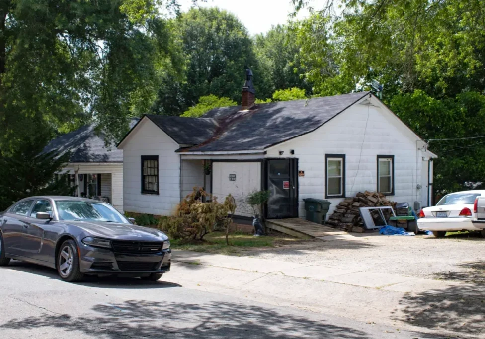 A white single-story house with a dark roof, surrounded by trees. A car is parked in front, and several items are scattered around the front yard and driveway.