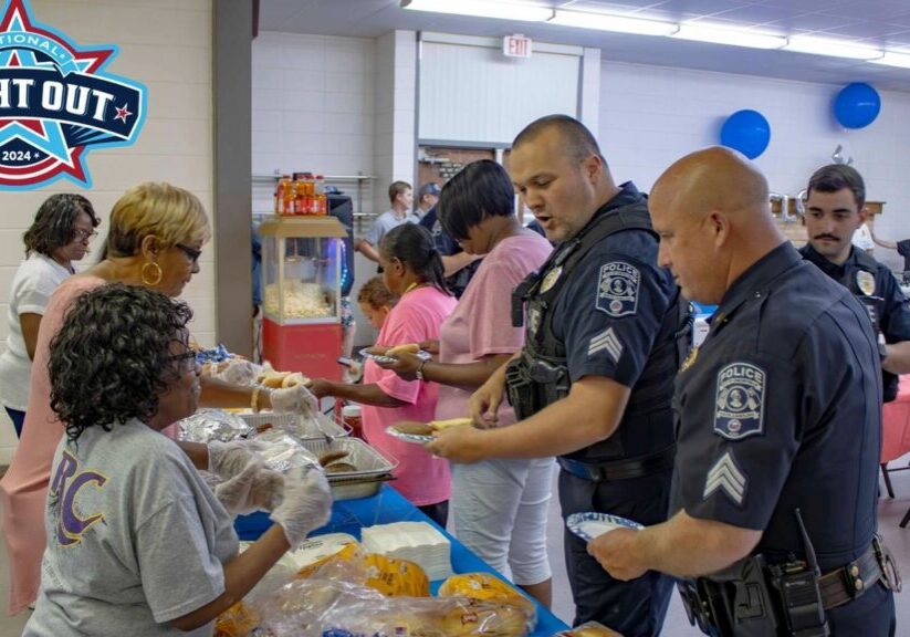 Police officers and community members gather in a room for a meal during the National Night Out event, with food being served by volunteers at a buffet table. National Night Out logo is displayed.