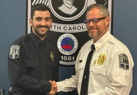 Two police officers shake hands in front of a sign that reads "City of Cherryville, North Carolina." Both are wearing uniforms, the officer on the left in black and the officer on the right in white.