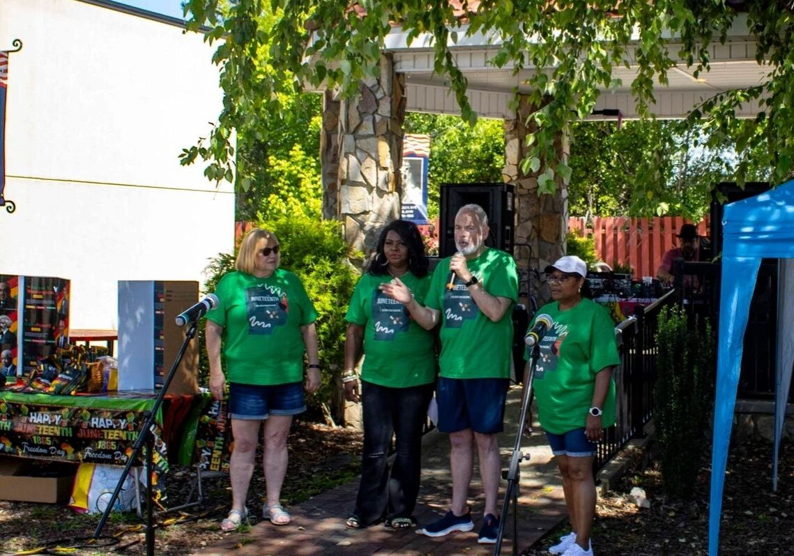 Four people wearing green shirts stand together on an outdoor stage under a tree, speaking into microphones. A small crowd is visible in the background.