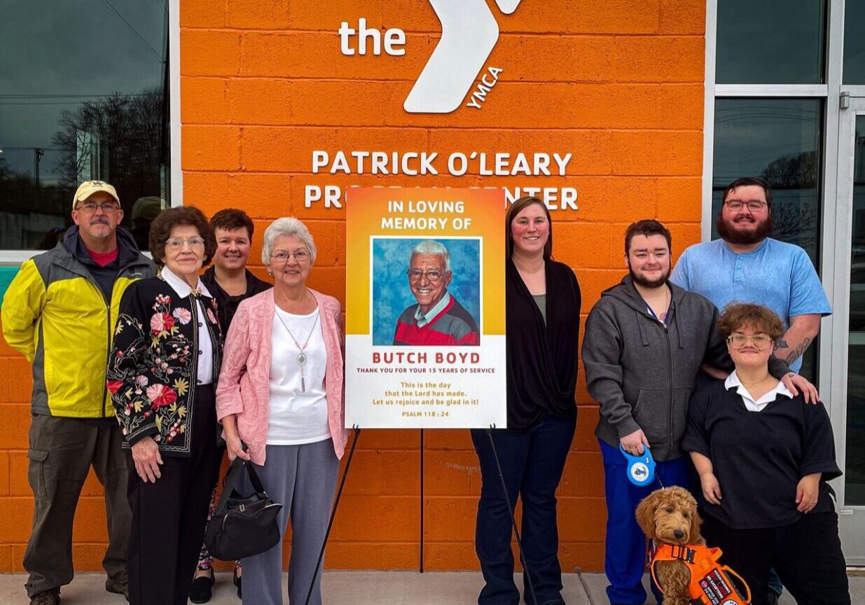 A group of people standing outside the Patrick O’Leary Prentis Center next to a poster in memory of Butch Boyd. One person holds a service dog wearing an orange vest.