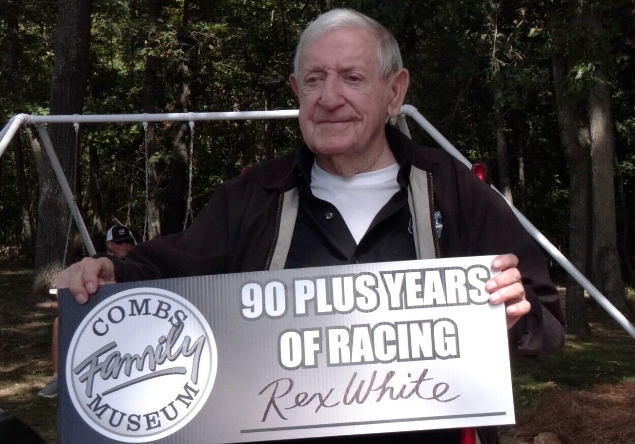 An elderly man holds a sign reading "90 Plus Years of Racing - Rex White" at the Combs Family Museum, with trees and a swing set in the background.