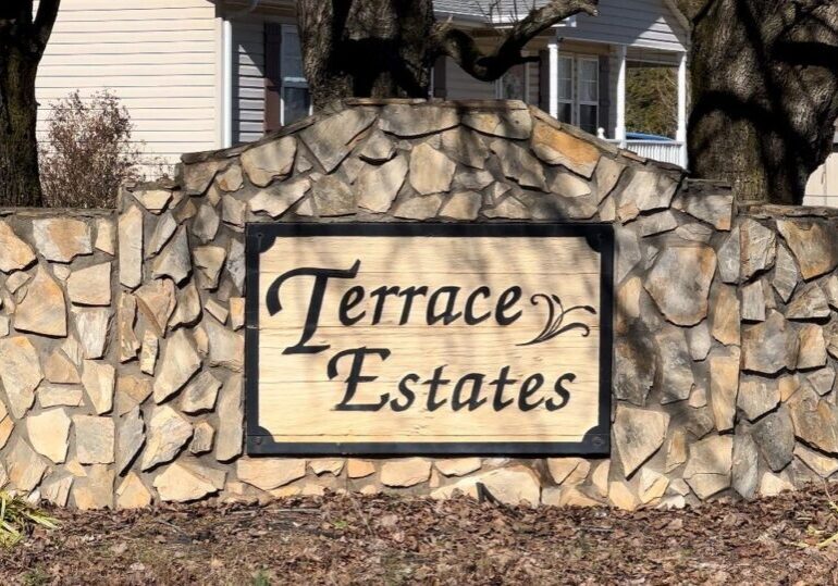 A large wooden sign with "Terrace Estates" engraved on it, mounted on a stone wall in front of a house.