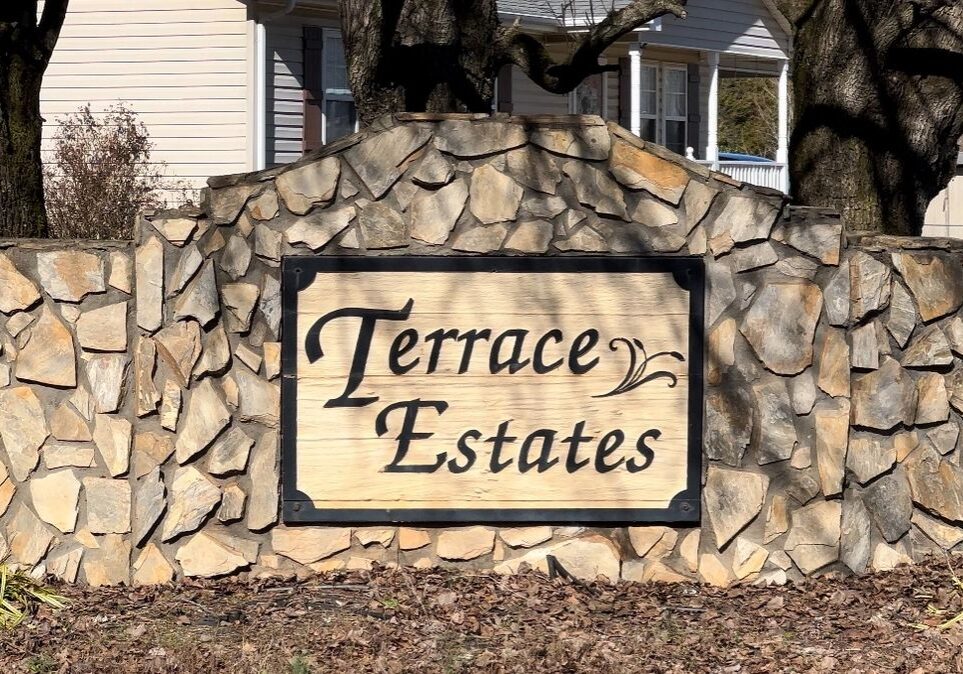 A large wooden sign with "Terrace Estates" engraved on it, mounted on a stone wall in front of a house.