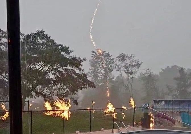 A bolt of lightning is striking near a swimming pool surrounded by trees and a metal fence during a storm, with mist and rain visible in the background.