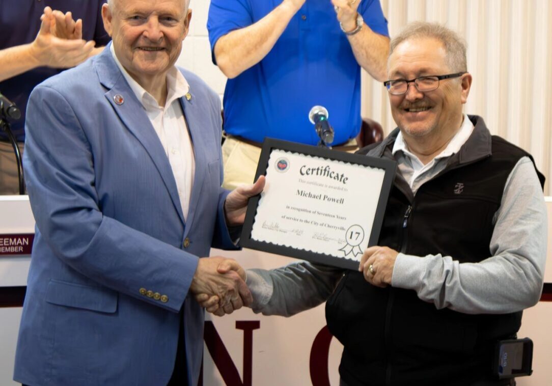 Two men are shaking hands. One man is holding a certificate while others in the background are clapping.