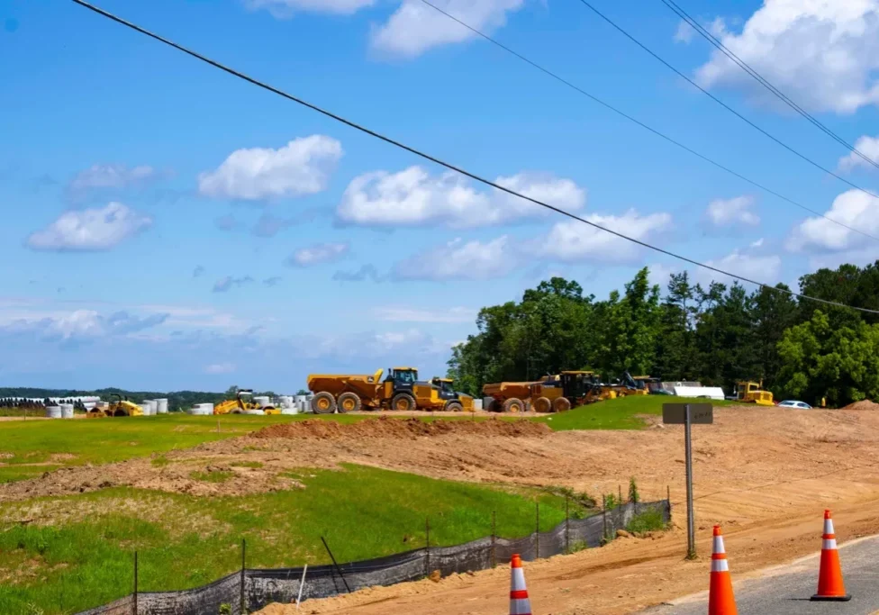 A construction site with heavy machinery, dirt piles, and a green field. Power lines cross above, and a traffic cone is placed near the edge of the road. Trees are visible in the background.