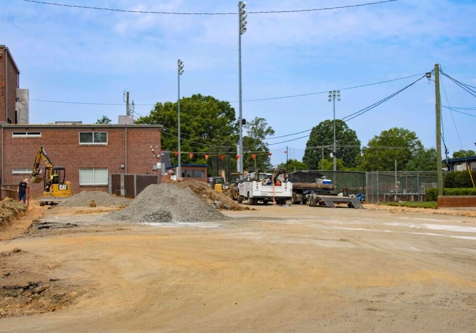 A construction site with heavy machinery and piles of gravel, adjacent to a brick building and a sports field with a chain-link fence. Trees and street lamps are visible in the background.