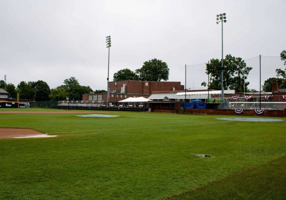 A baseball field with a grassy outfield and a brick building in the background, featuring bleachers, light poles, and bunting decorations.