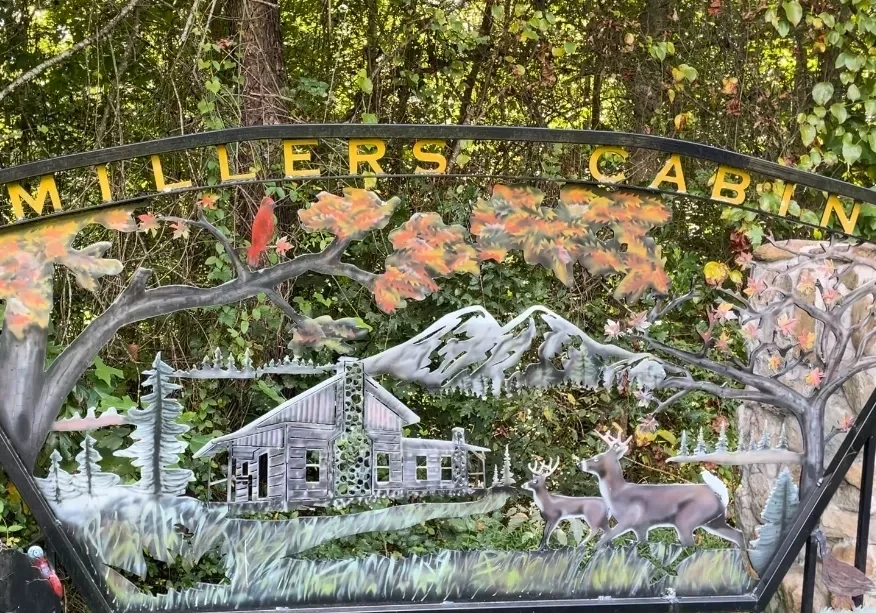 A metal sign featuring a cabin in the woods with mountains in the background and two deer in the foreground. The text "Millers Cabin" arches above the scene.