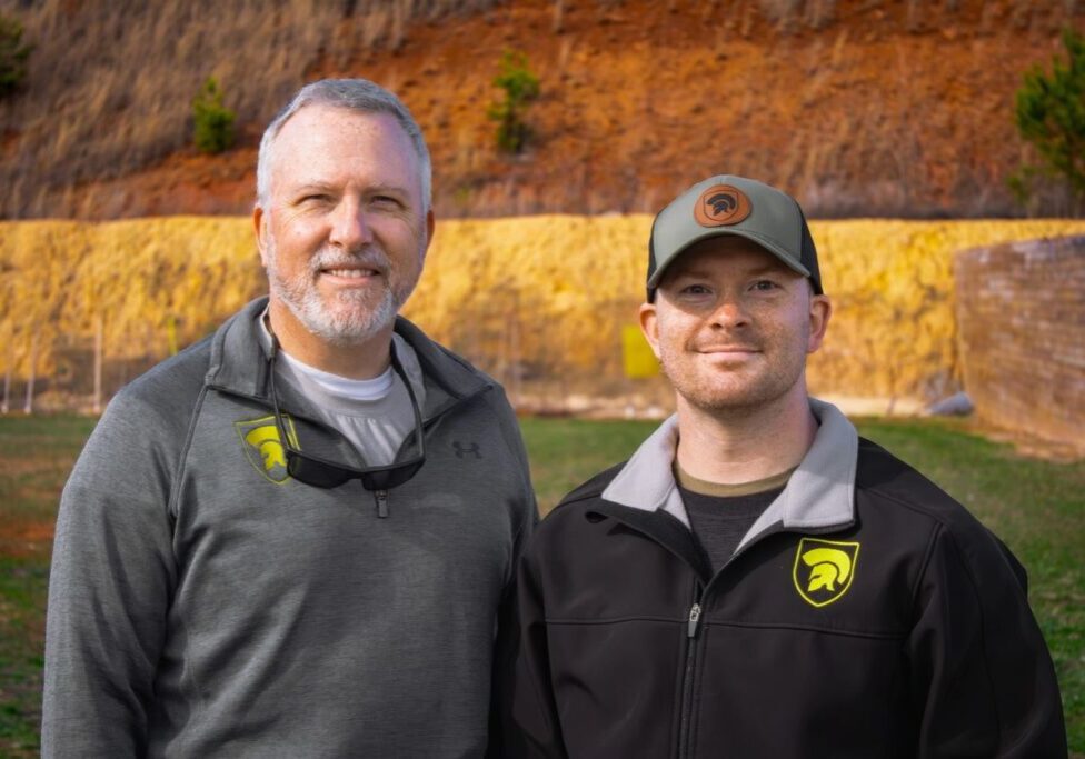 Two men wearing matching outdoor gear with bear logo stand smiling in front of a wooded hillside and a yellow barrier.