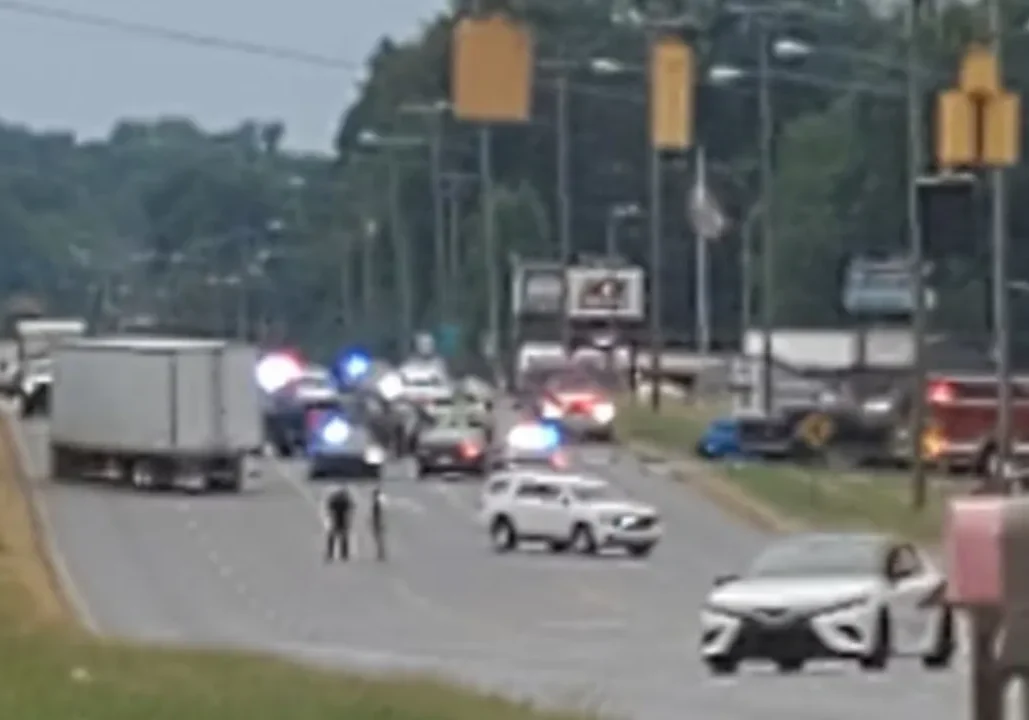 A road with multiple police vehicles and flashing lights blocking traffic. A large truck and several cars are also on the road. Some officers are standing near the vehicles.