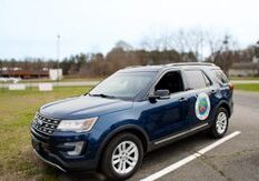 A blue SUV with a logo on the front door is parked in a parking lot with a grassy area and trees in the background.